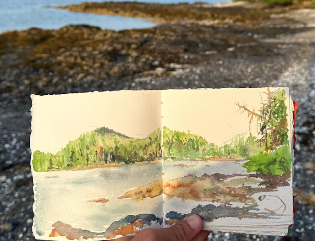 Close up of a hand holding a sketchbook with a painting in front of the same landscape with water, trees, and distant mountains