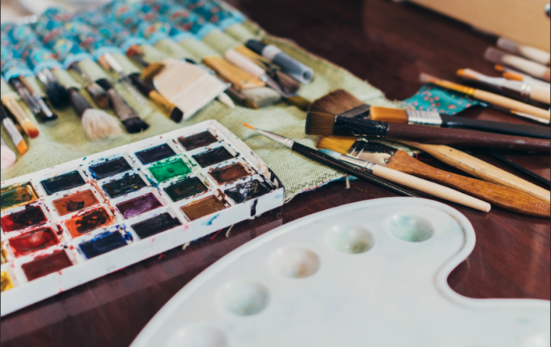 Watercolor paints, brushes, and a pallete sitting on top of a wooden table