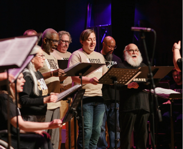Veterans Choir performing at the Spring 2025 showcase at the Seattle Art Museum. Singers standing in a row with music stands in front of them, all singing.