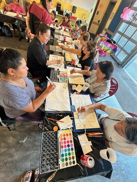 Picture of artists seated around a table painting with watercolors on paper.